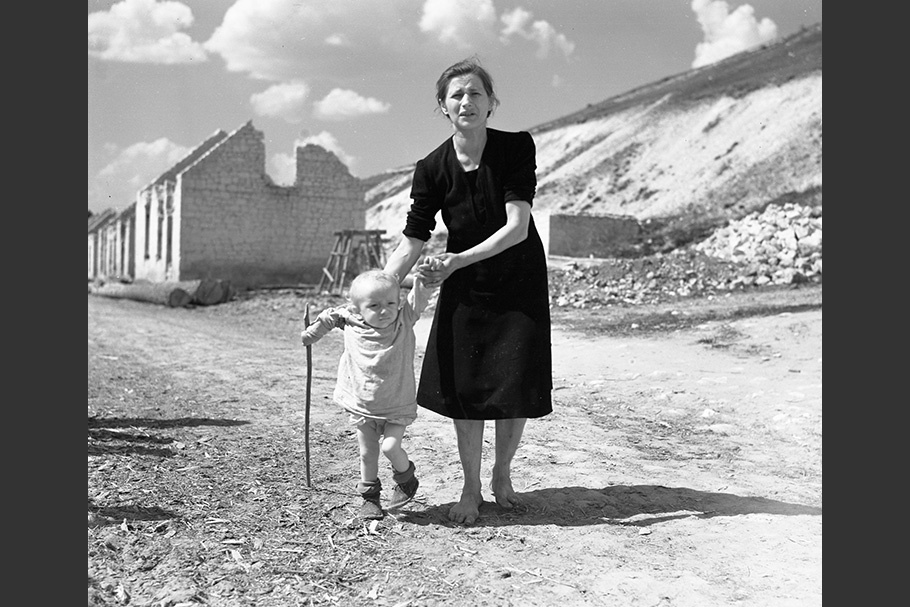 A mother and child walk down a ruined street.