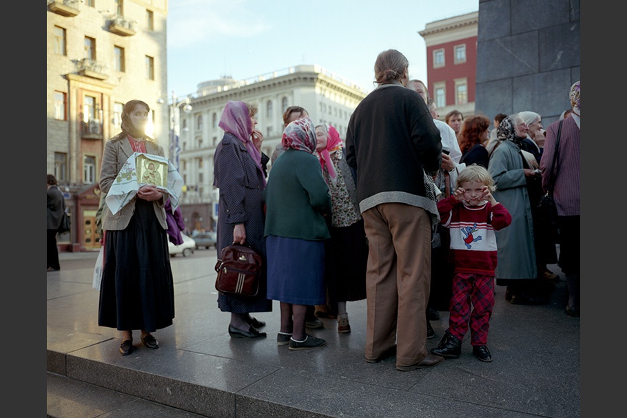 A crowd of people on the street, including a child making a face and a woman holding an icon. 