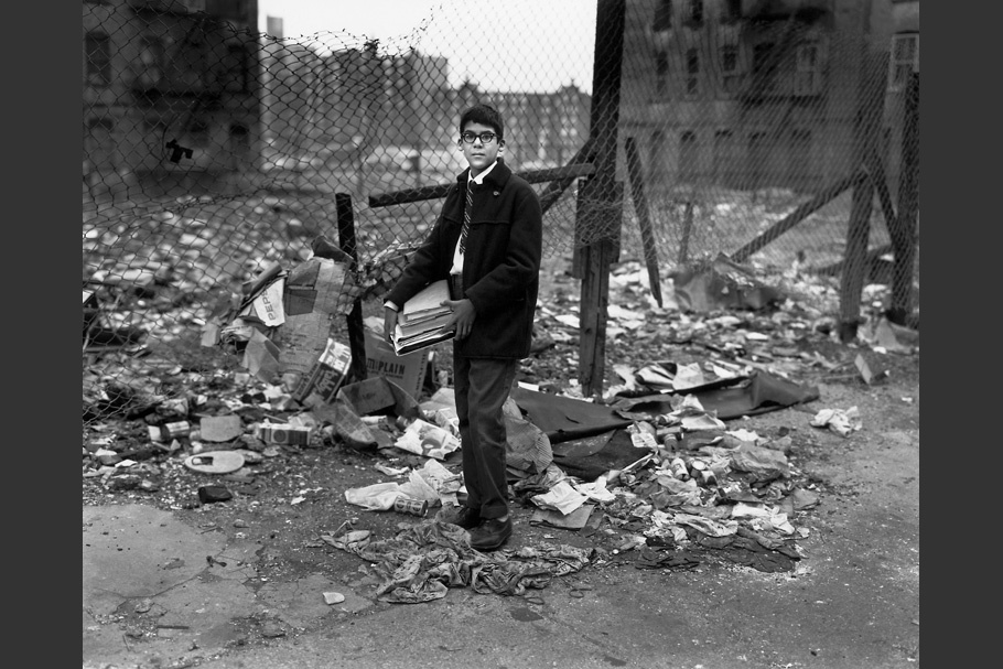 A boy carrying books past a vacant lot.