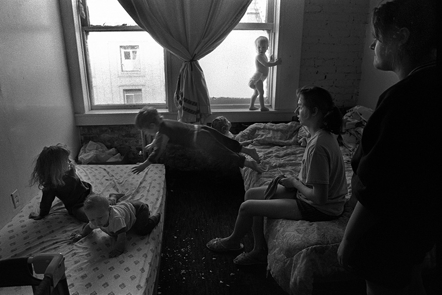 Women and children playing in a bedroom, with a child jumping between beds.
