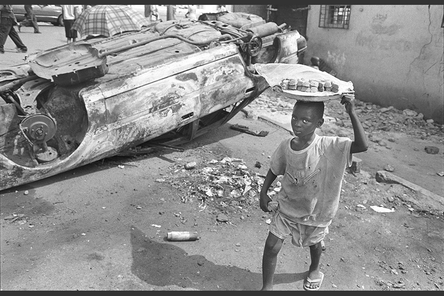 A boy walking by an overturned car carrying a tray on his head.