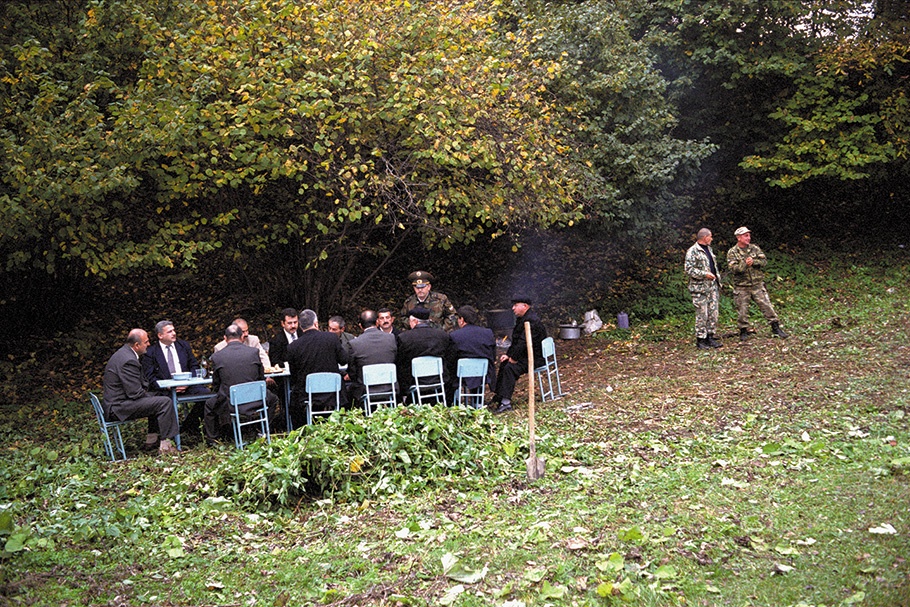Men in suits seated at an outdoor table. 