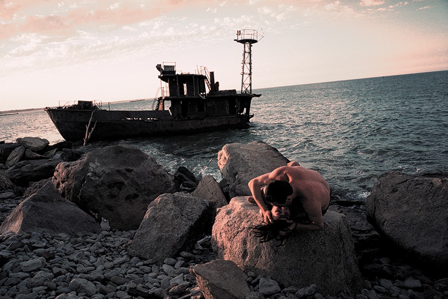 A couple embraces on rocks in front of a boat.