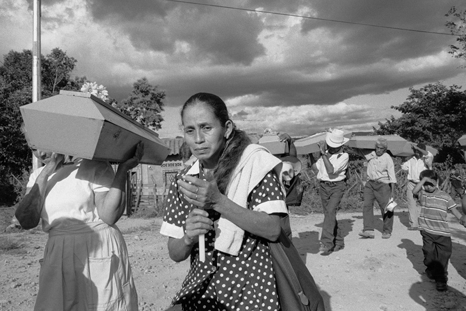 A woman in a polka dot dress walking with a procession of coffins.