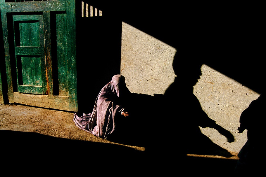 A woman begging in front of a sun and shadow streaked wall.