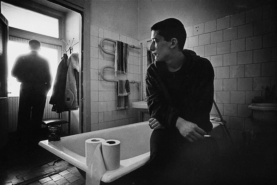 A man sitting on a bathtub and another looking out a window.