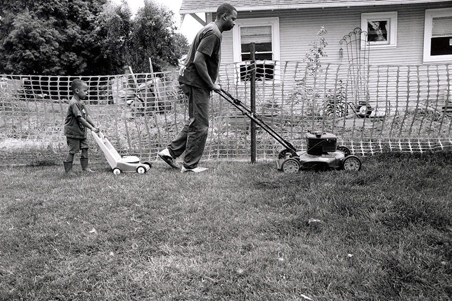 A man with a lawnmower and a boy with a toy lawnmower.