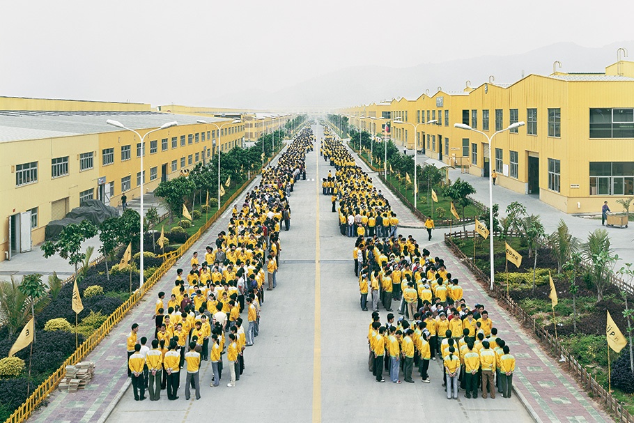 Workers in yellow standing in formation on a street surrounded by yellow factories.