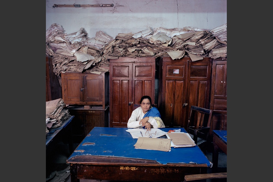 Piles of paper behind woman at desk.