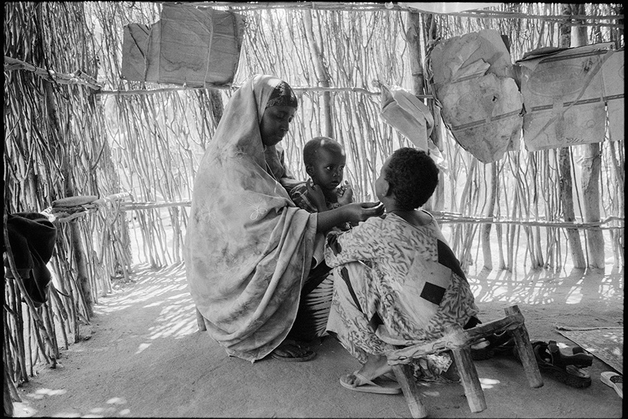 Woman with two children in front of a wall made of sticks.