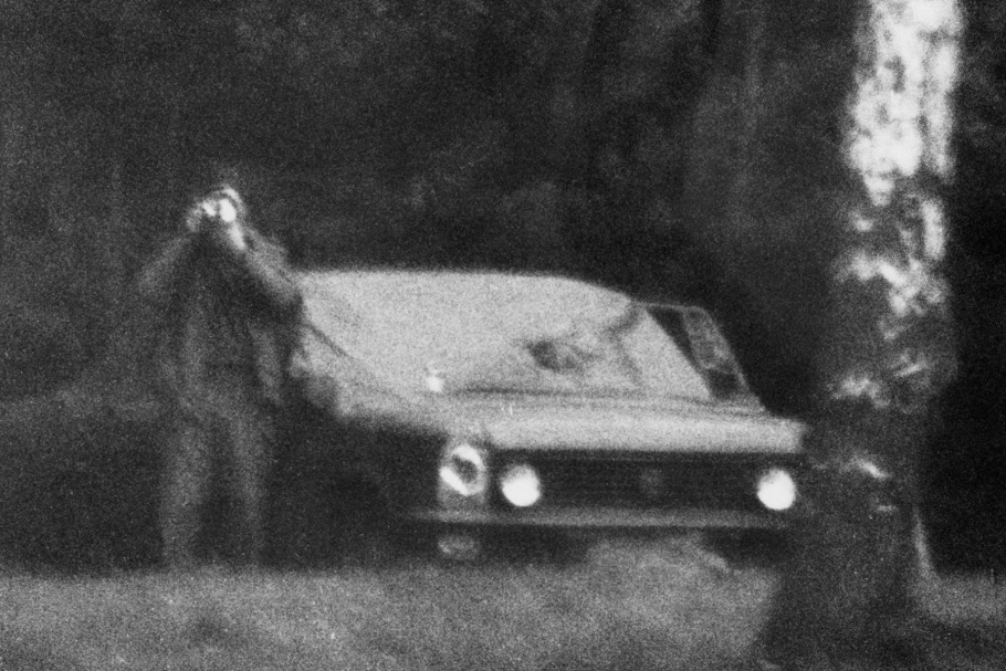 Man standing to the left of a parked car holding camera to his face