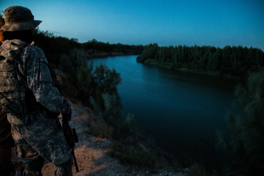 A man wearing camouflage and holding a rifle stands near a river