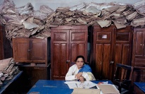 Piles of paper behind woman at desk.